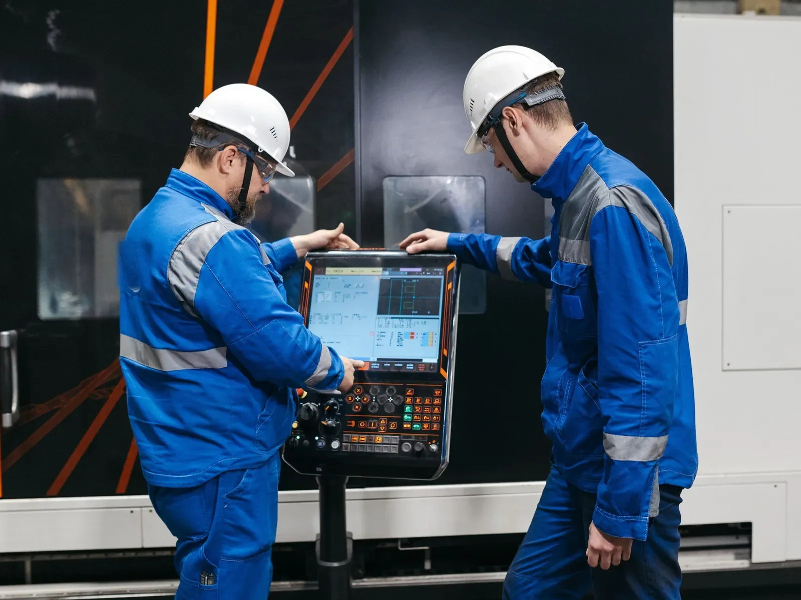 A senior engineer shows a younger colleague the controls of a machine on a factory floor